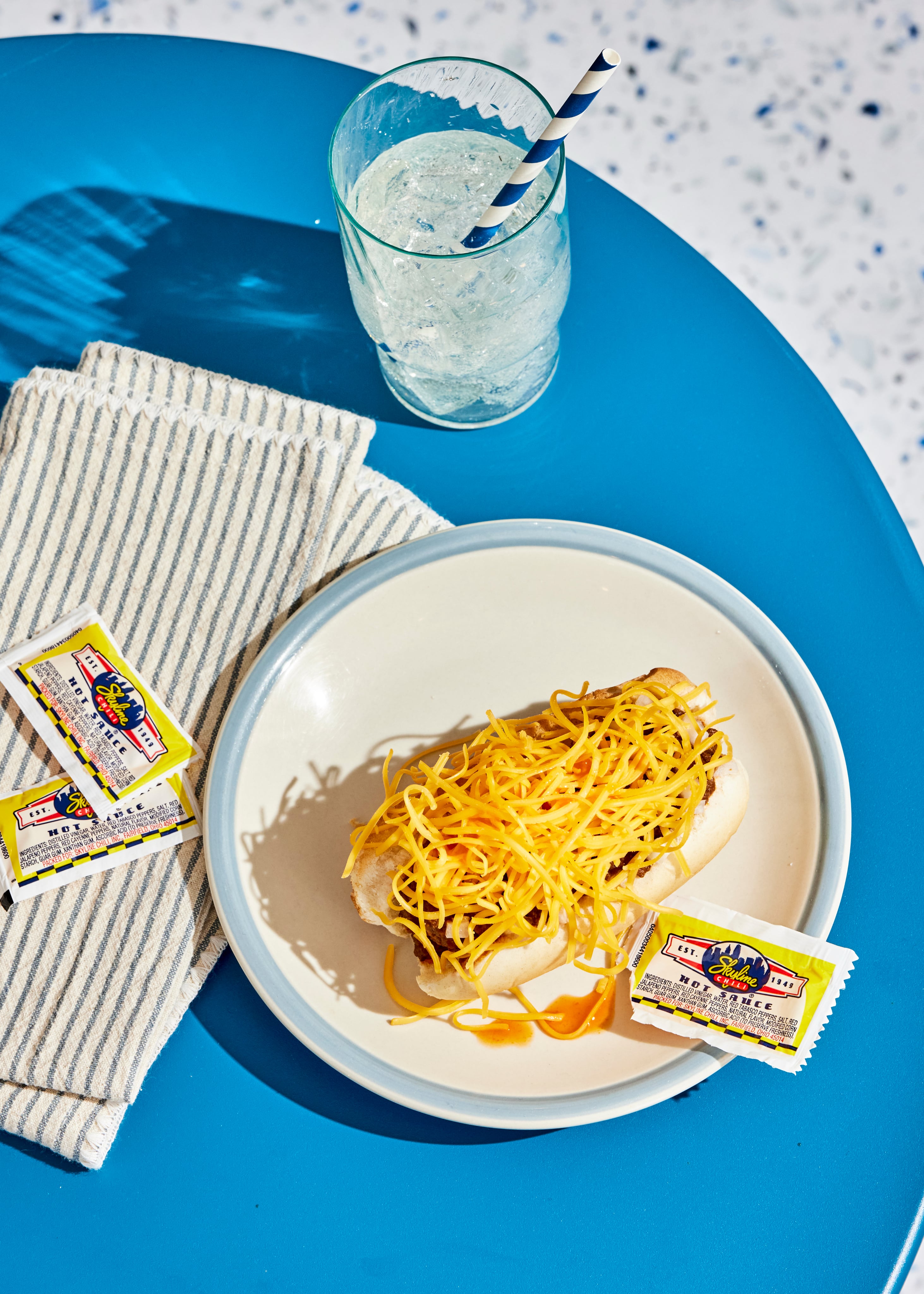 Blue table with plate with a coney on it, 3 sauce packets surrounding it and a striped napkin and glass of water